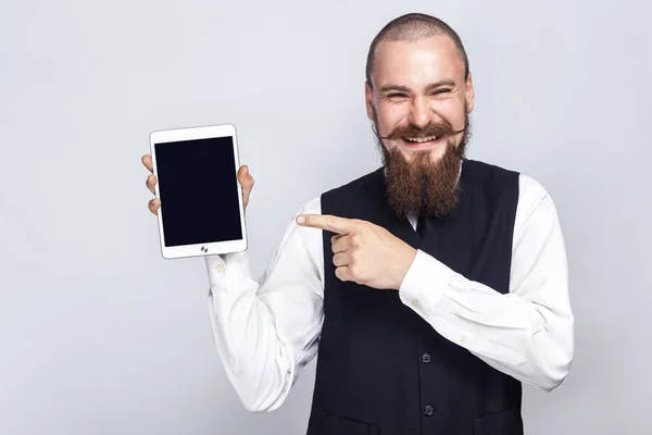Stilig affärsman med skägg och mustasch håller digital tablett och tittar på kameran och visar skärmen med fingret och skrattar. Studio skott, på grå bakgrund. — Stockfoto