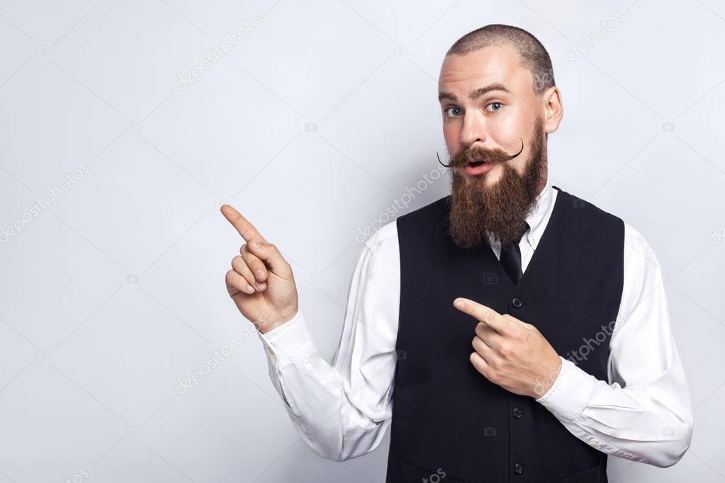 Handsome businessman with beard and handlebar mustache looking at camera, surprised and showing copy space with fingers. studio shot, on gray background.