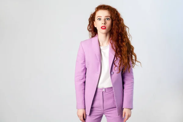 Portret van verrast mooie zakenvrouw met rood - bruin haar en make-up in roze pak. Kijkend naar camera, studio schoot op grijze achtergrond. — Stockfoto