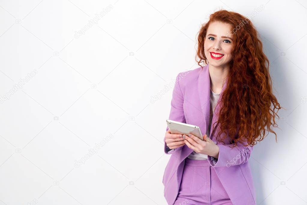Portrait of successful happy beautiful business woman with red - brown hair and makeup in pink suit holding tablet, looking at camera with toothy smile. studio shot on gray background.
