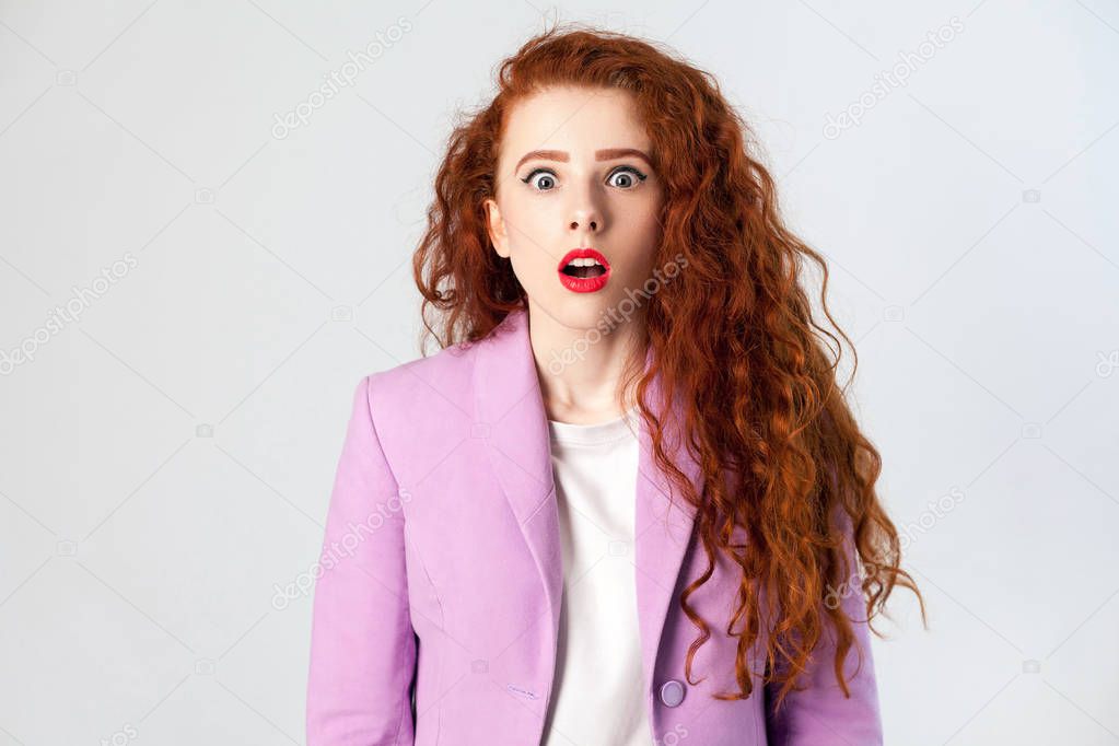 Portrait of shocked beautiful business woman with red - brown hair and makeup in pink suit. looking at camera, studio shot on gray background.