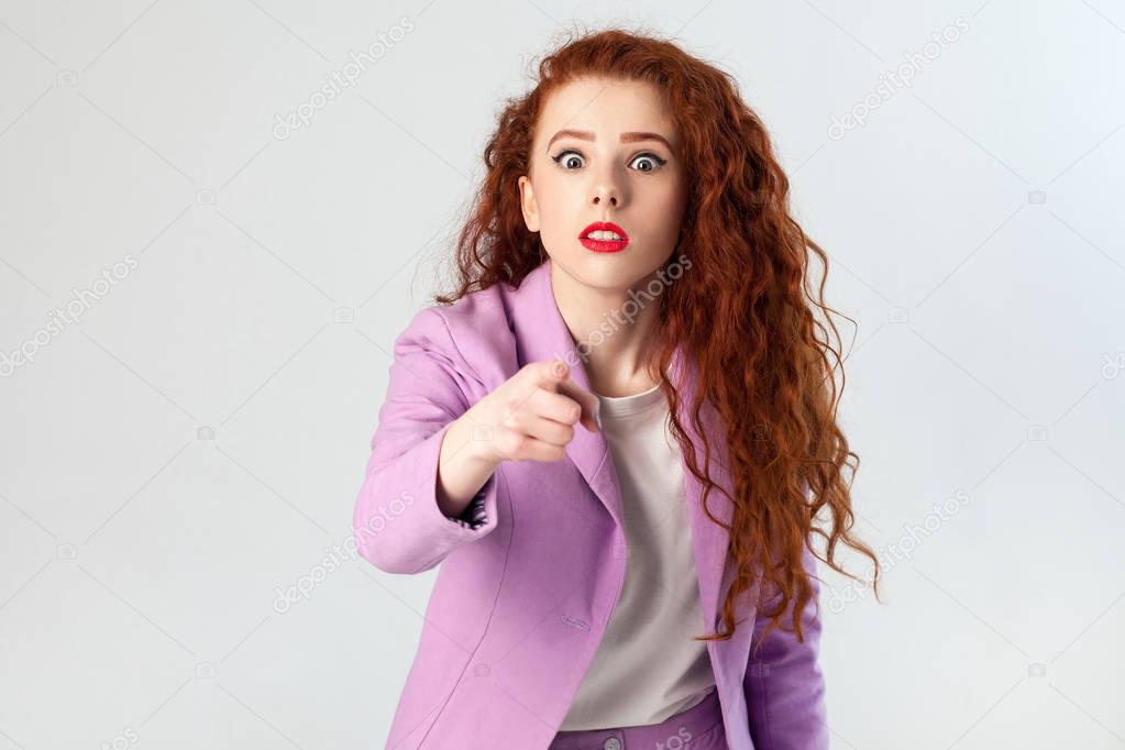Portrait of shocked beautiful business woman with red - brown hair and makeup in pink suit. pointing and looking at camera, studio shot on gray background.