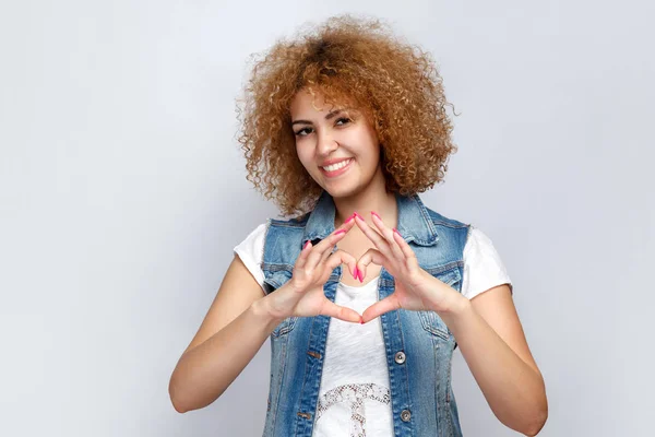 Ritratto di bella ragazza di razza mista riccia in stile casual con forma di cuore con le mani guardando la fotocamera. studio girato su sfondo grigio chiaro . — Foto Stock