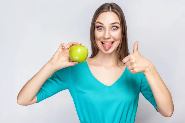 Joven hermosa mujer con pecas y vestido verde sosteniendo manzana con los pulgares hacia arriba y la lengua . — Foto de Stock