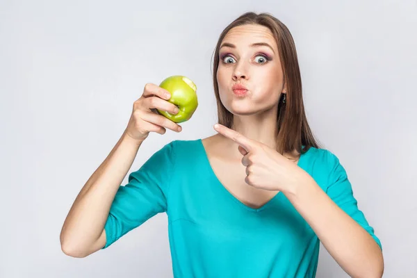 Young beautiful woman with freckles and green dress holding and eating apple and pointing and looking at camera with big eyes. — Stock Photo, Image