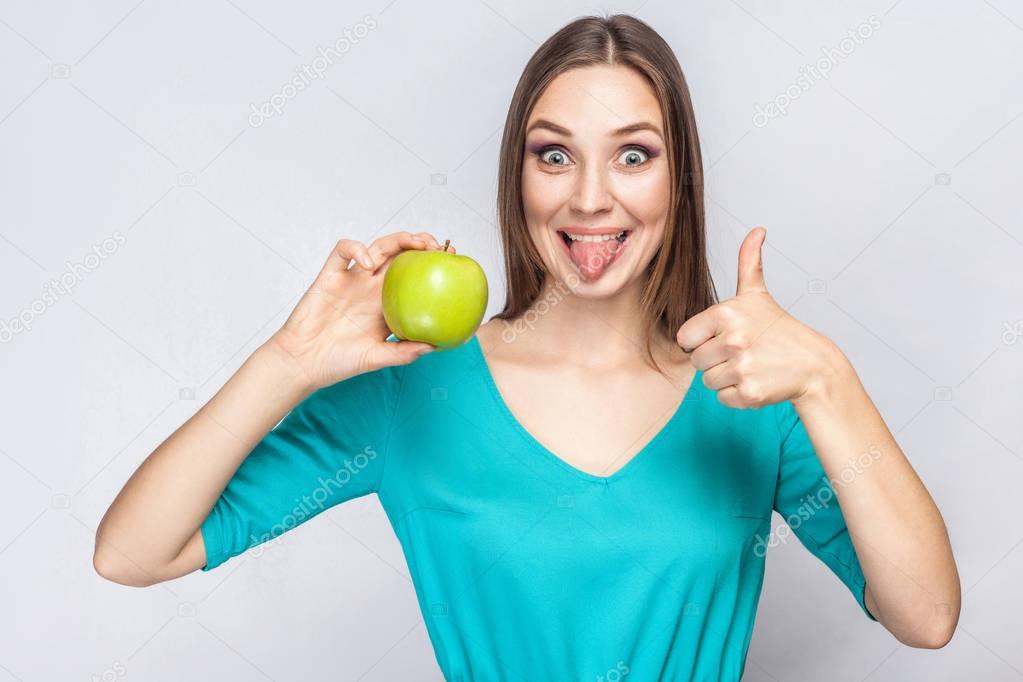 Young beautiful woman with freckles and green dress holding apple with thumbs up and tongue. 