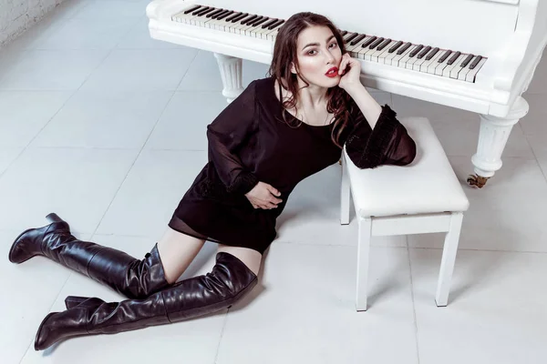 Модель в черном платье и сапогах с дымчатым макияжем глаз и прической сидит на полу и позирует рядом с пианино. студия съемки . — стоковое фото