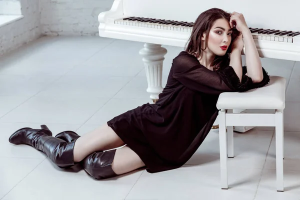 Modelo de moda con vestido negro y botas con ojos ahumados maquillaje y peinado sentado en el suelo y posando cerca con piano. plano de estudio . — Foto de Stock