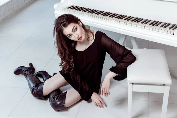 时装模特与黑色的连衣裙和靴子的烟熏眼妆的化妆和发型坐在地板上和附近和钢琴的合影。工作室拍摄. — 图库照片
