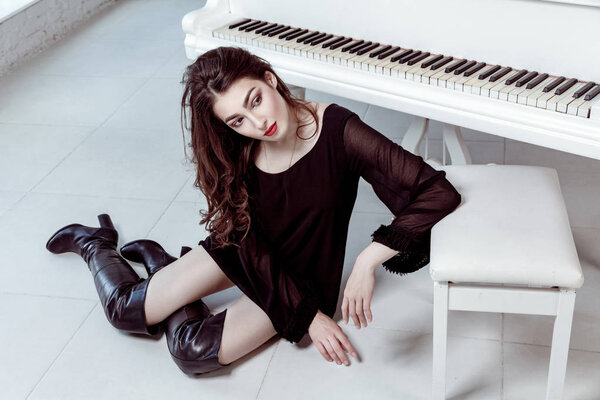 Fashion model with black dress and boots with smokey eyes makeup and hairstyle sitting on floor and posing near with piano. studio shot.