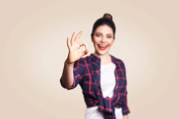 - Хорошо. Счастливая зубастая улыбающаяся молодая женщина показывает знак ОК с пальцами. студийная съемка на бежевом фоне. фокус на руке . — стоковое фото