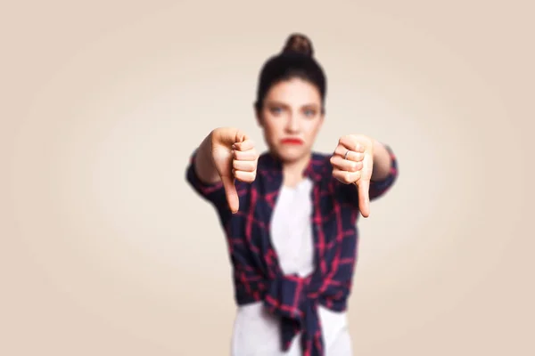 N'aime pas. Jeune fille contrariée malheureuse avec un style décontracté et les pouces de cheveux de chignon vers le bas de son doigt, sur le mur blanc beige avec l'espace de copie regardant la caméra avec le sourire de dent. se concentrer sur le doigt . — Photo