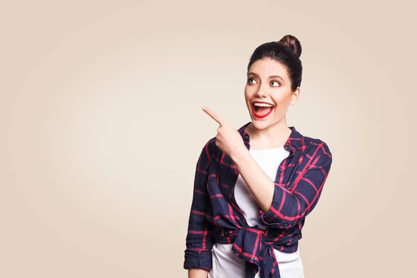 Joven chica feliz con estilo casual y pelo moño apuntando con el dedo hacia los lados, demostrando algo en la pared en blanco beige con espacio de copia para su información o contenido promocional . —  Fotos de Stock