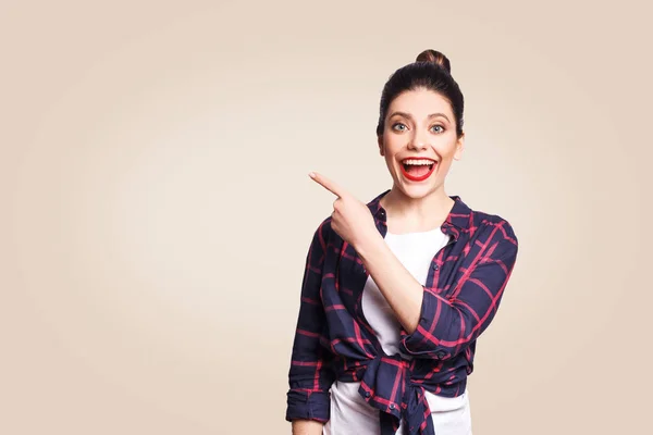 Young happy girl with casual style and bun hair pointing her finger sideways, demonstrating something on beige blank wall with copy space for your information or promotional content. — Stock Photo, Image