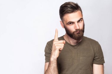 Portrait of serious bearded man with warning finger and dark green t shirt against light gray background. studio shot.  clipart