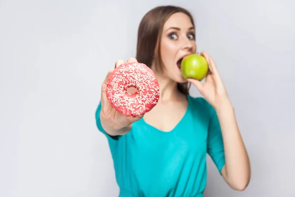 Beautiful young woman with freckles in green dress, eating green apple and holding pink donut. studio shot on light gray background — Stock Photo, Image