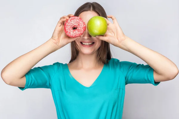 Mulher bonita com sardas em vestido verde, segurando diante de seus olhos maçã verde e donut rosa com sorriso de dente. estúdio filmado em fundo cinza claro . — Fotografia de Stock