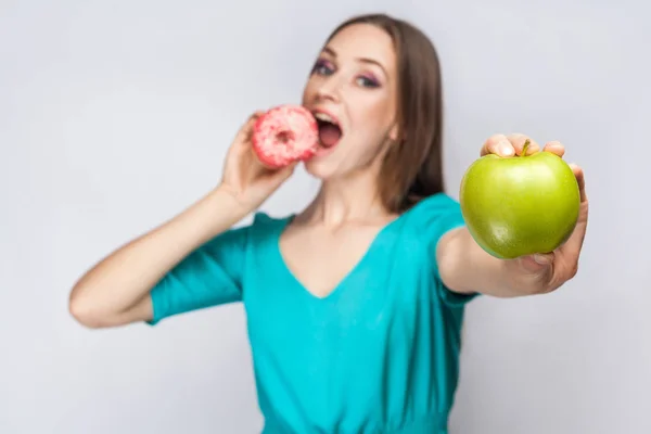 Beautiful young woman with freckles in green dress, eating pink donut and holding green apple. studio shot on light gray background — Stock Photo, Image