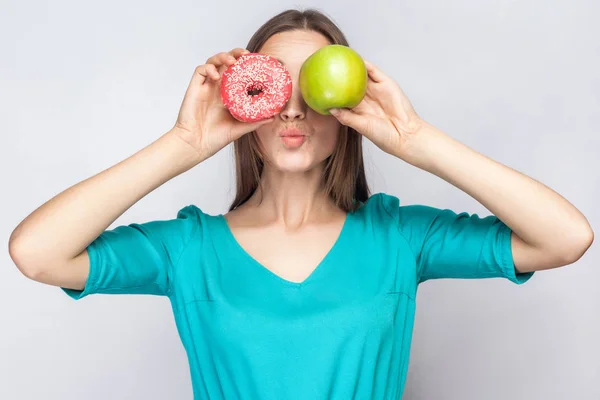 Beautiful young woman with freckles in green dress, holding before her eyes green apple and pink donut and kissing. studio shot on light gray background. — Stock Photo, Image