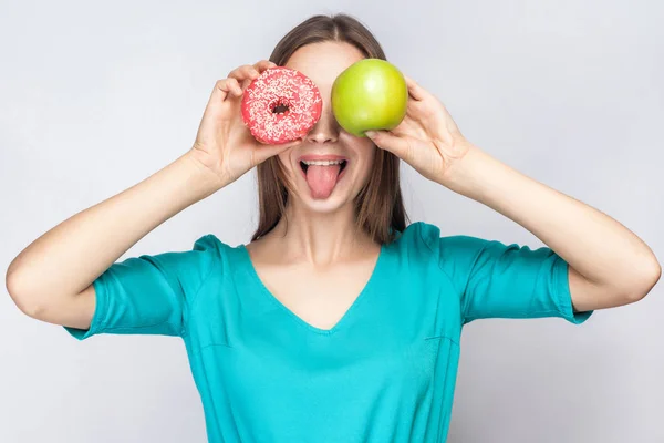 Красива молода жінка з веснянками в зеленому платті, тримає перед очима зелене яблуко і рожевий пончик і показує язик. студійний знімок на світло-сірому фоні . — стокове фото