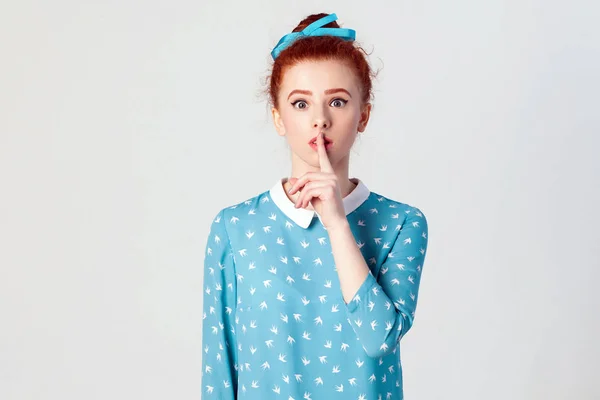 La hermosa joven pelirroja, sosteniendo el dedo índice en los labios, levantando las cejas, diciendo "Shh", "Silencio", "Tsss". Estudio aislado sobre fondo gris — Foto de Stock