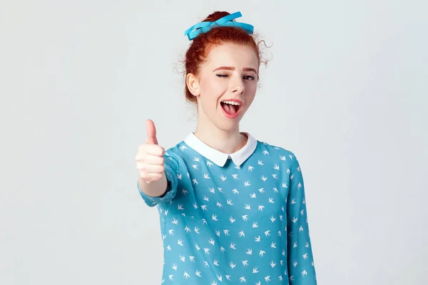 A menina ruiva alegre bonita nova que mostra o polegar acima e o sorriso do dente e pisca na câmera. Estúdio isolado filmado em fundo cinza — Fotografia de Stock