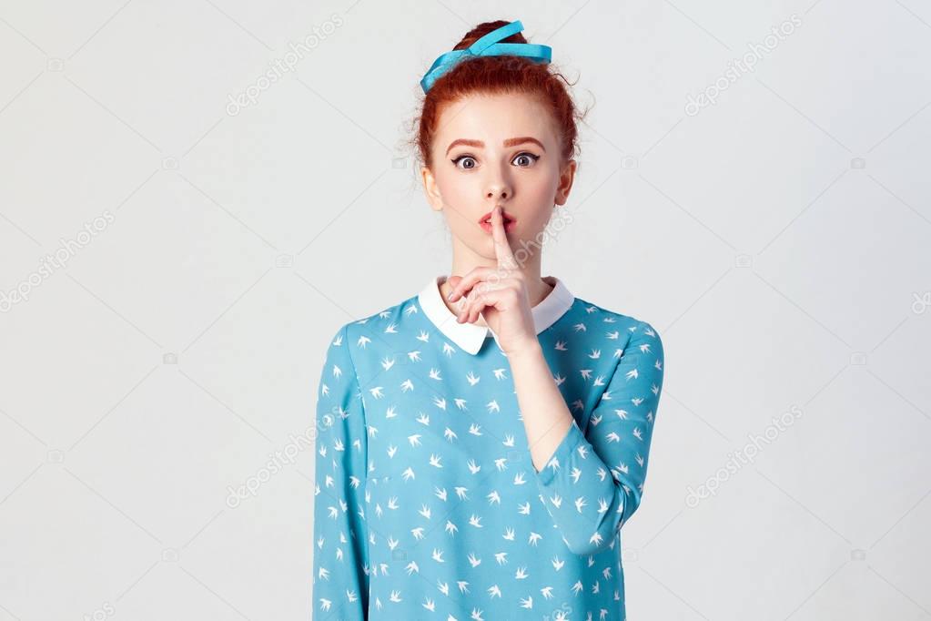 The beautiful young redhead girl, holding index finger at lips, raising brows, saying 