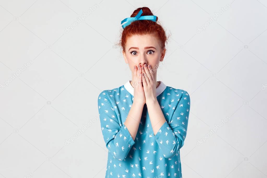 Portrait of serious young ginger. Caucasian girl covering mouth with both hands keeping a secret. Beautiful redhead female in blue dress doesn't want to spread rumors or some confidential information. Isolated studio shot on gray background.