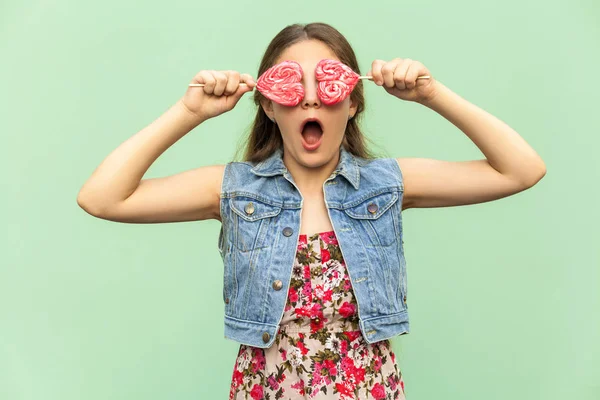 Het tiener meisje met lang haar van blondie, in geschokt, bezit twee lolipop op haar ogen, grappig kijken. Geïsoleerde studio opname op lichtgroene achtergrond. — Stockfoto