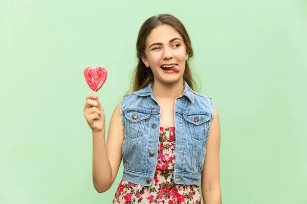 Joven adolescente con el pelo largo rubio, guiño a la cámara, lengua hacia fuera, sostiene piruleta, teniendo mirada divertida. Estudio aislado sobre fondo verde claro . — Foto de Stock