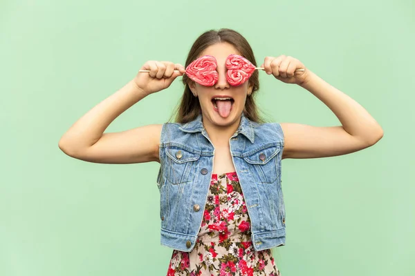 Mooie tiener meisje met lang haar van blondie, op zoek op camera en tong uit, houdt twee lolipop, grappig kijken. Geïsoleerde studio opname op lichtgroene achtergrond. — Stockfoto