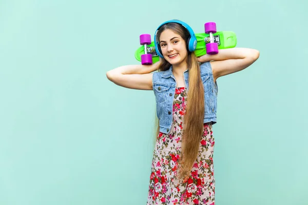 Šťastné krásné dlouhé vlasy blond dívka v šatech a sluchátka, baví se zelenou plastovou penny skateboard v přední části světle modré zdi. Izolované studio zastřelil — Stock fotografie