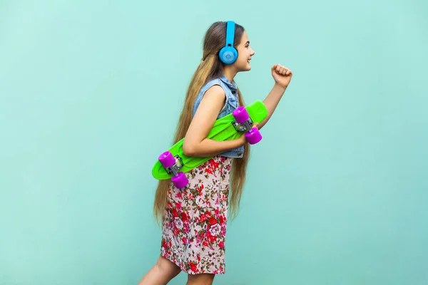 Hipster in motion on light blue background. The young happy beautiful long haired blonde girl in dress and headphones, holding green plastic penny skateboard. Isolated studio shot — Stock Photo, Image