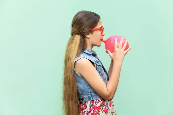 Jeune fille heureuse soufflant des ballons pour la fête d'anniversaire, regardant drôle à la caméra sur un fond vert clair. tournage studio intérieur — Photo