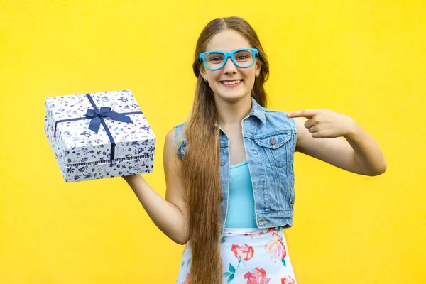 Das schöne lächelnde Mädchen in Kleid und blauer Brille, Zeigefinger auf Geschenkbox und zahmes Lächeln, isoliert über gelbem Hintergrund. Indoor-Studiodreh — Stockfoto