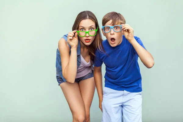 Eyewear concept. WOW faces. Young sister and brother with freckles on their faces, wearing trendy glasses, posing over light green background together. Looking at camera with surprised face. Studio shot — Stock Photo, Image