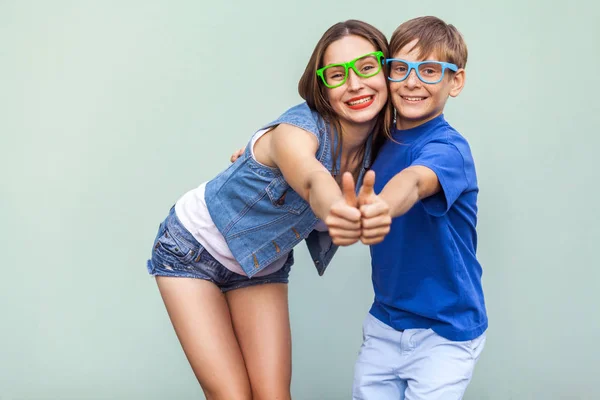 Familie, Emotionen und Gefühle. Die ältere Schwester und ihr Bruder mit Sommersprossen posieren gemeinsam auf blauem Grund und blicken mit einem zahmen Lächeln und hochgereckten Daumen in die Kamera. Studioaufnahme — Stockfoto