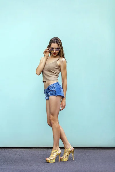 Modelo de moda sexy en gafas, jeans cortos, top de estilo casual y zapatos dorados, poses sensuales sobre el fondo azul claro. Tocando su cara. Interior, plano de estudio —  Fotos de Stock