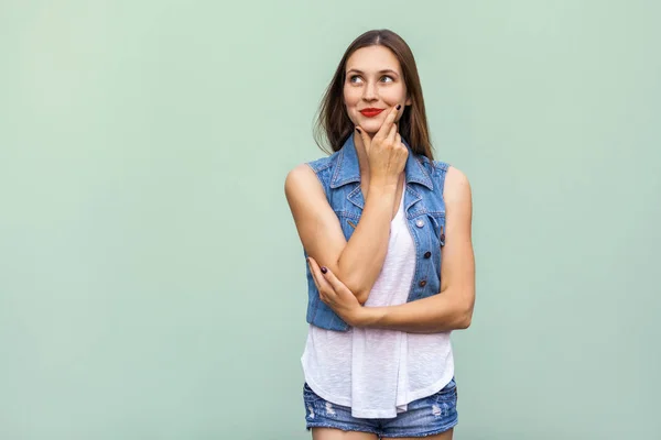 Happy cheerful teenage girl with freckles, casual style white t shirt and jeans jacket, looking up, thinking and touching her face and a, enjoying good day and free time indoors. Studio shot, light green background — Stock Photo, Image