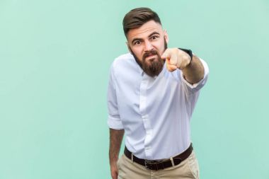 Angry bearded businessman with white shirt looking at camera and pointing finger to you. Studio shot isolated on light green background. clipart