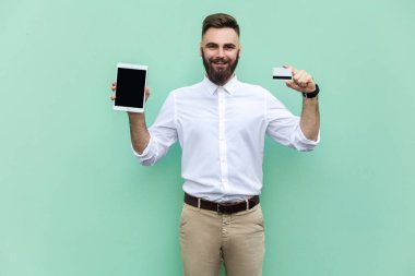 Online alışveriş veya bankacılık. Eller kredi kartı ve tablet tutarak ve kamera ve dişlek gülümseme bakarak genç yetişkin iş adamı. Kapalı, stüdyo vurdu. Açık yeşil arka planı üzerinde izole