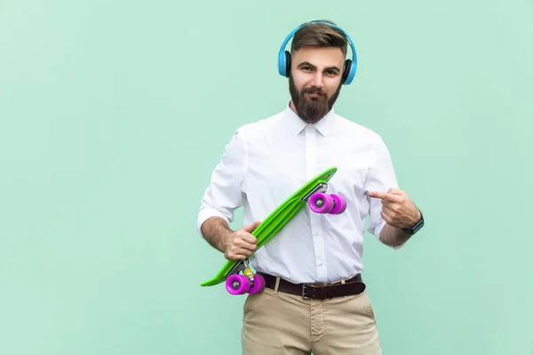 Hombre joven empinado con auriculares azules y brazos de penique monopatín. Fondo verde claro. Captura de estudio — Foto de Stock