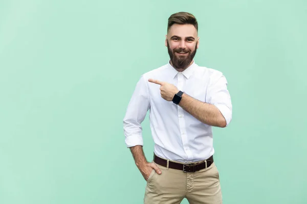 Zakenman aanwijsapparaat kopie ruimte. Knappe jonge volwassen man met baard in wit overhemd camera te kijken en weg te wijzen terwijl staande geïsoleerd op lichtgroene achtergrond — Stockfoto