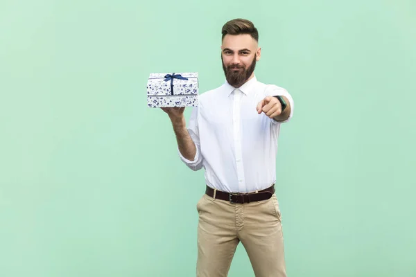Young adult bearded man, pointing at camera and holding a gift box. Studio shot on light green background — Stock Photo, Image