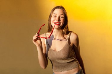 Flört. Muhteşem genç çilli modeli Güneş gözlüğü kamera ve dişlek gülümseme bakarak. Sarı arka plan üzerinde izole. Stüdyo vurdu