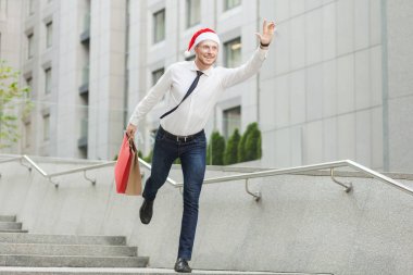 Young adult bearded man in santa hat holding many shopping bags and gifts and running to kids. clipart