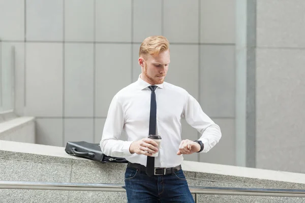 Homme d'affaires aux cheveux rouges regardant la montre intelligente et tenant tasse . — Photo