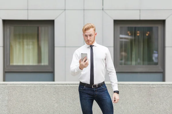 Cabeza roja gerente adulto joven mirando el teléfono inteligente con la cara conmocionada . — Foto de Stock