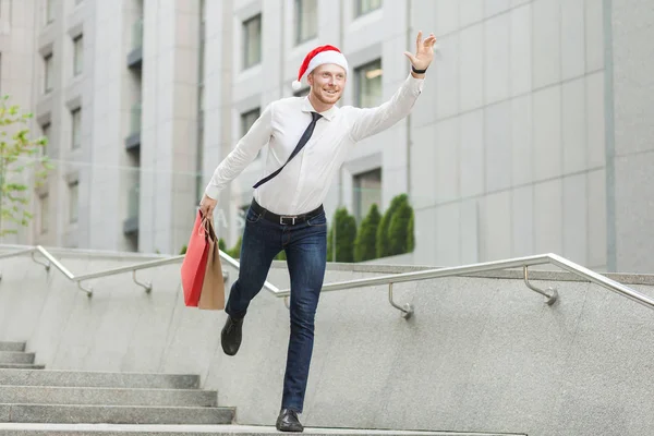 Junger erwachsener bärtiger Mann mit Weihnachtsmütze hält viele Einkaufstüten und Geschenke in der Hand und rennt zu Kindern. — Stockfoto