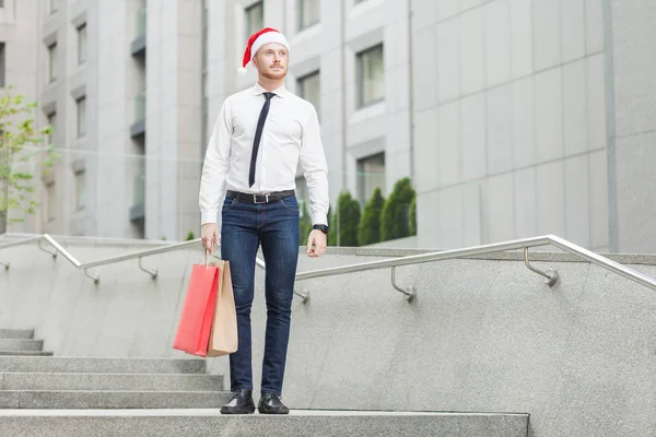 Éxito joven adulto jengibre hombre de negocios con sombrero rojo de santa, mirando hacia otro lado y sosteniendo regalos —  Fotos de Stock